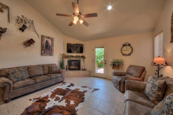 A living room with a brown leather sofa, a brown leather armchair, a fireplace, a ceiling fan, and a cowhide rug.