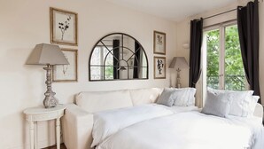 A bedroom with a large bed, a white sofa, a small table with a lamp, and a large window with curtains. - Amazing Balcony and Pinch-Yourself Eiffel Views! (Paris)