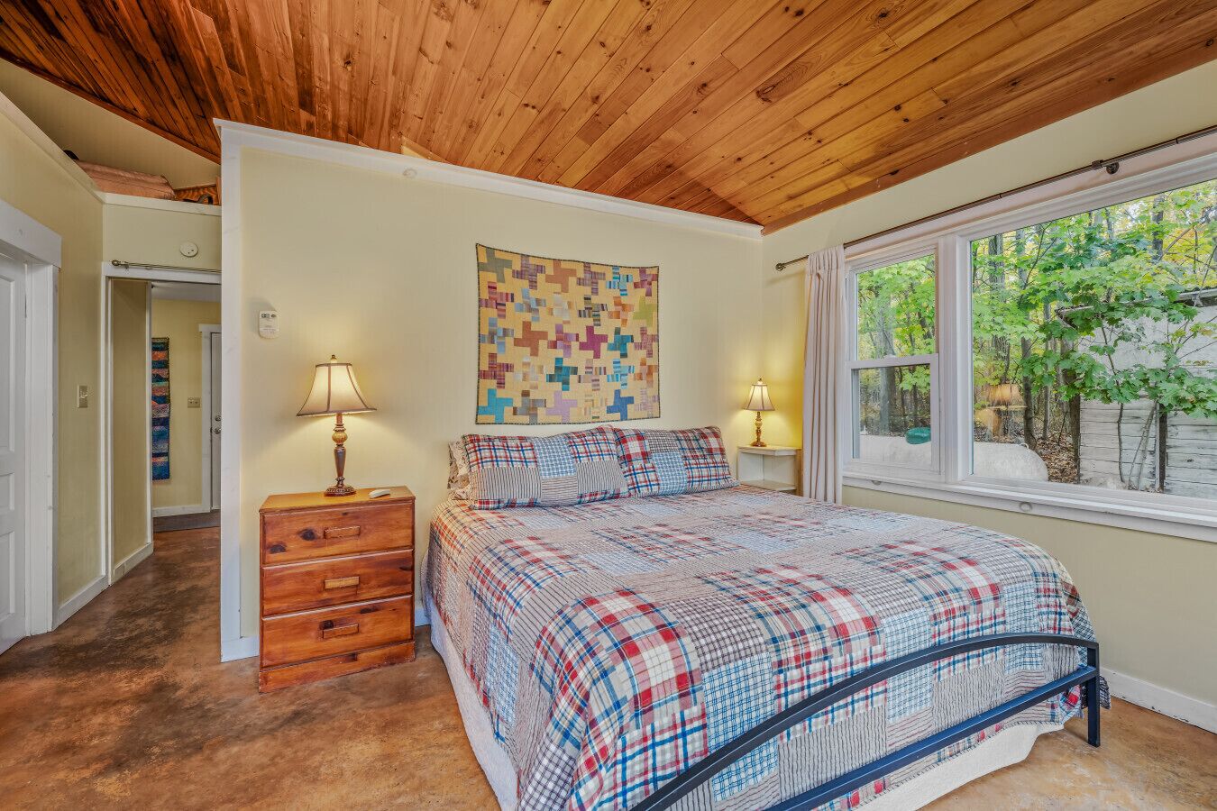 A bedroom with a wooden ceiling, a bed with a plaid bedspread, two bedside lamps, a wooden nightstand, a quilted wall hanging, and a window with a view of trees.