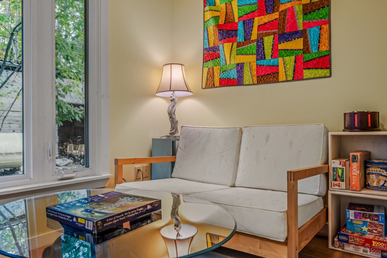 Un salon chaleureux avec un canapé, une table basse en verre, une lampe et une bibliothèque contenant des jeux de puzzle.