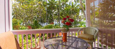 A covered outdoor seating area with a round table and two chairs, a potted plant with red flowers, and a view of palm trees.