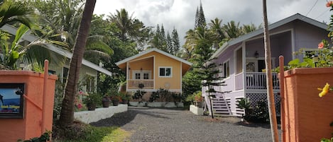 Uma cabana amarela com varanda, cercada por vegetação tropical e um caminho de cascalho.