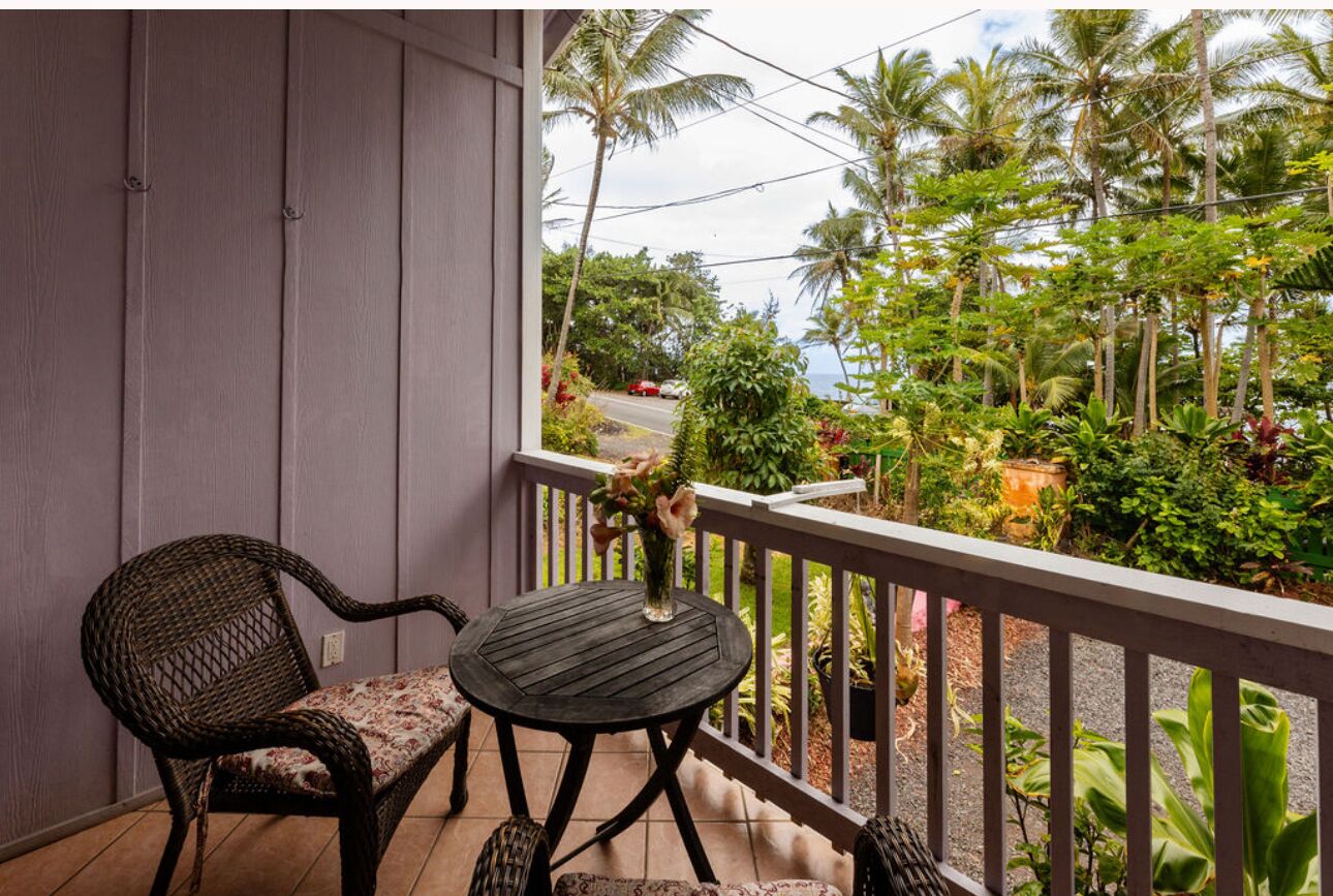 A balcony with wicker chairs and a small table, overlooking a lush garden and a road with a car.