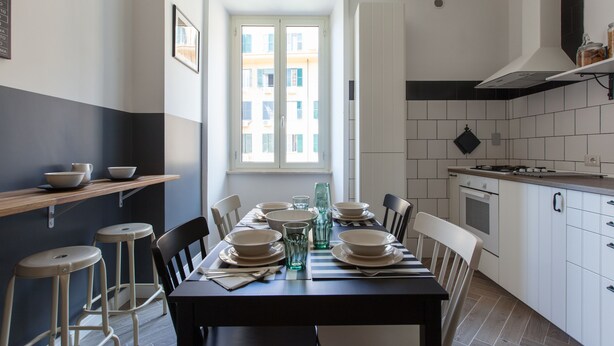 A modern kitchen with a dining area, featuring a large window, white cabinets, and a striped tablecloth. - Pannonia Smart House (Rome)