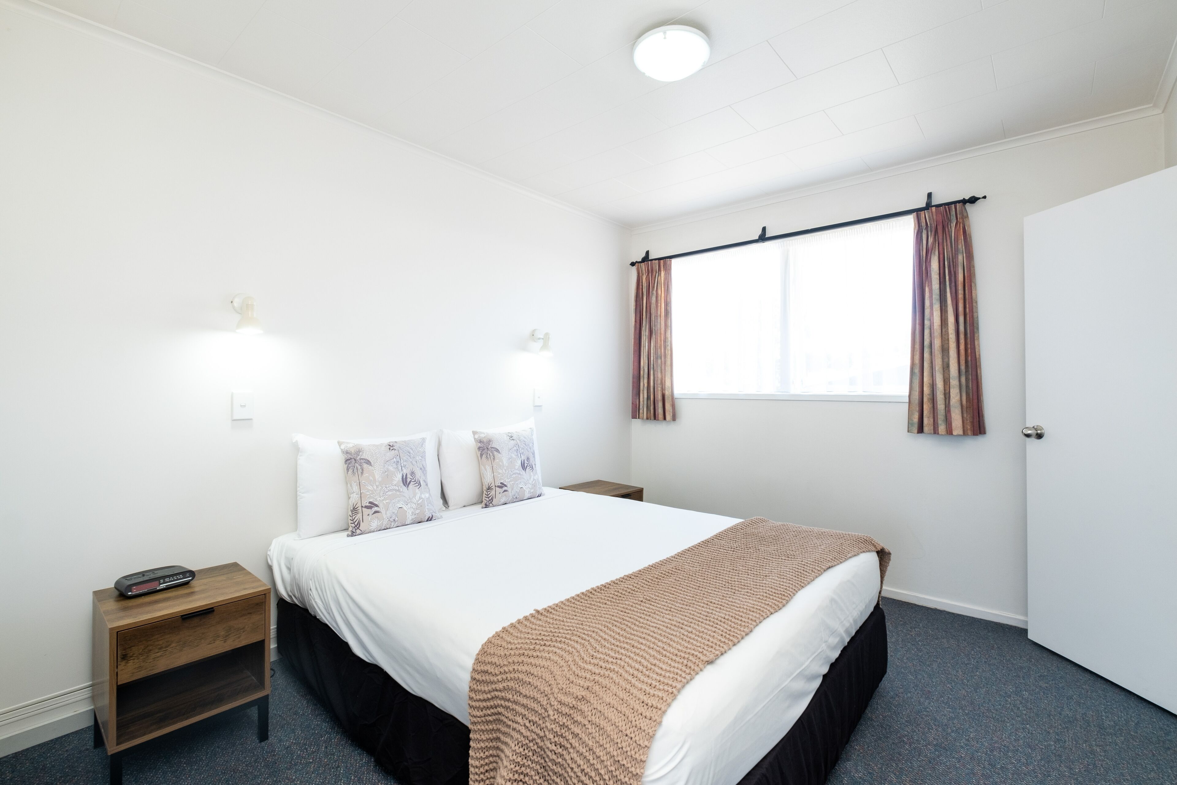 A hotel room with a bed, bedside table, lamp, and window with curtains.