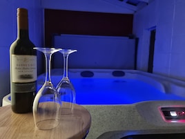 Una botella de Hawke's Bay Merlot Malbec Cabernet Sauvignon sobre una mesa de madera, con dos copas de vino a su lado. Se ve una bañera de hidromasaje al fondo.