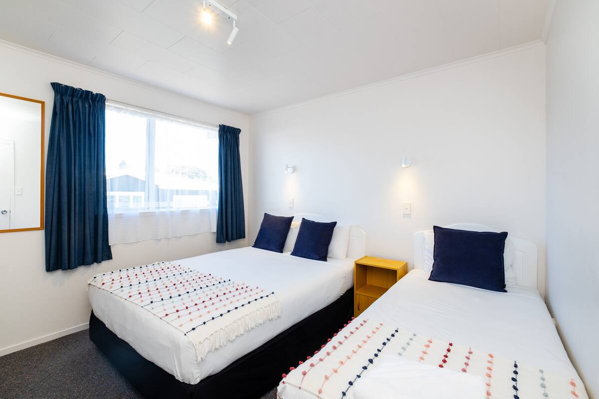 A hotel room with two beds, a window with curtains, a bedside table, and a wall-mounted light.