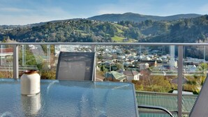 A balcony with a glass table and a chair, overlooking a town and hills. - Baldwin Street BNB (Dunedin)