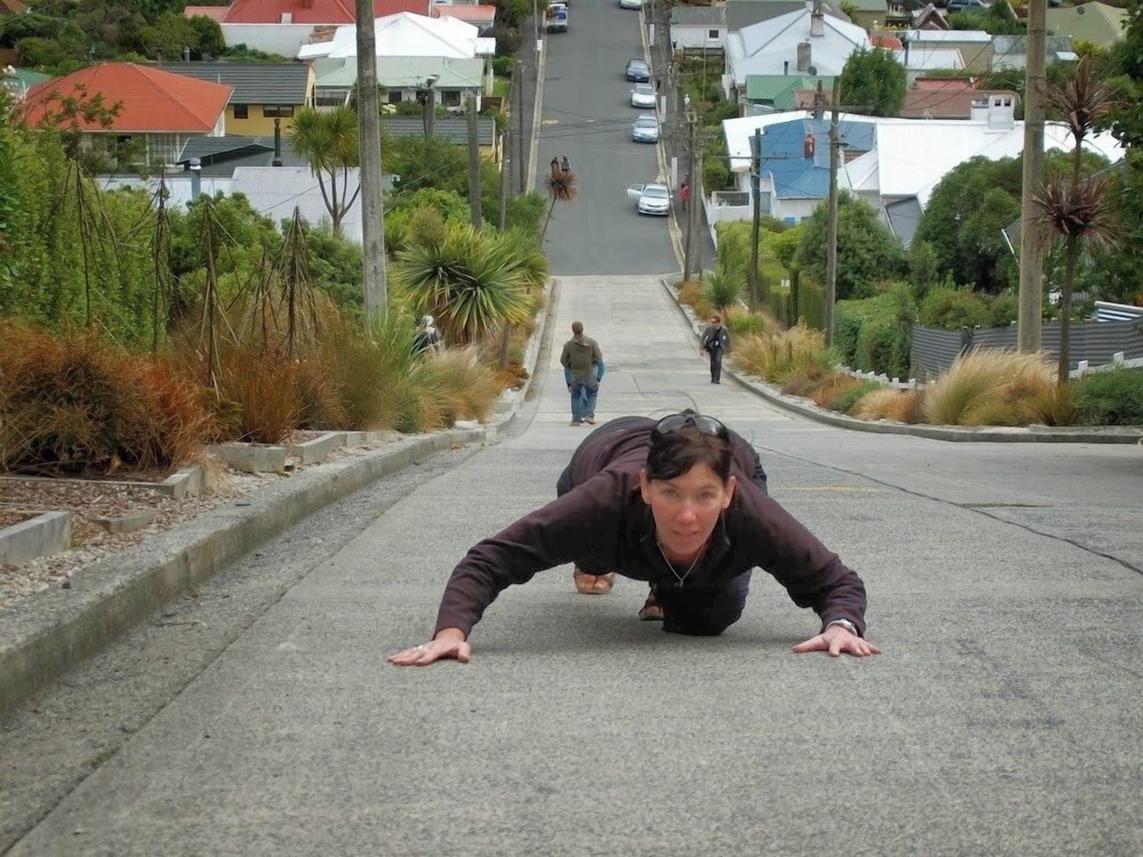 A person doing push-ups on a residential street with houses and greenery on both sides.