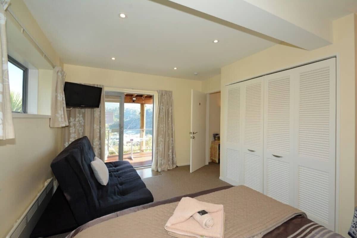 A bedroom with a bed, a massage chair, a TV, and a sliding glass door to a balcony.