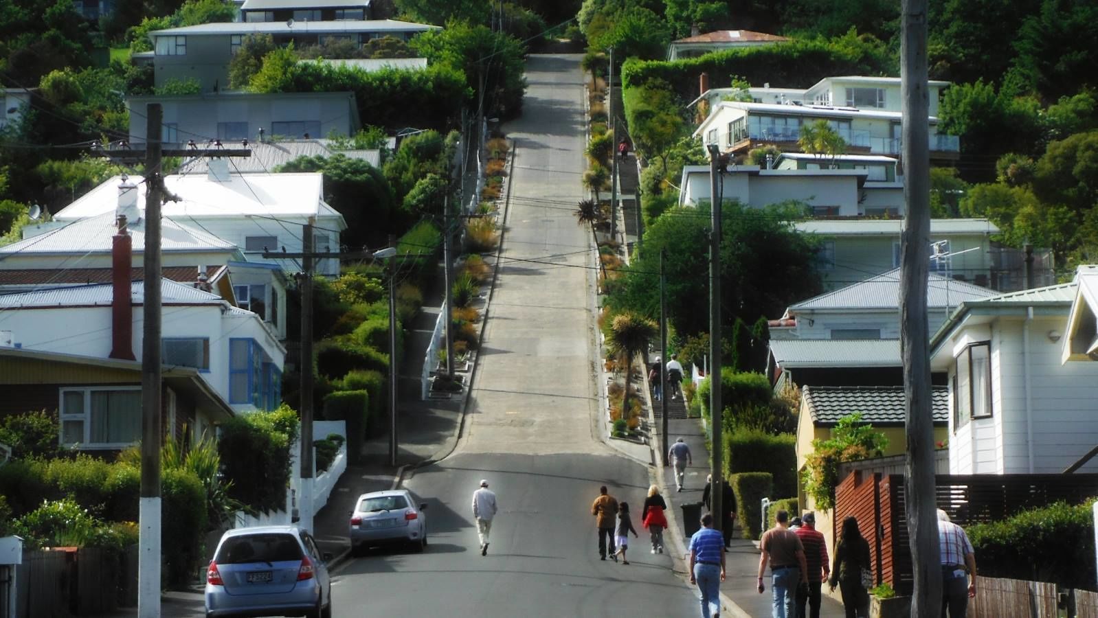 A residential street with houses, parked cars, and pedestrians.