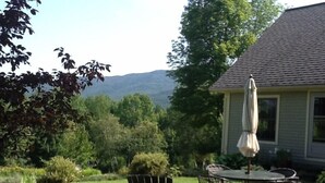 A patio with a table and chairs, a white umbrella, and a view of a house and mountains in the background.
