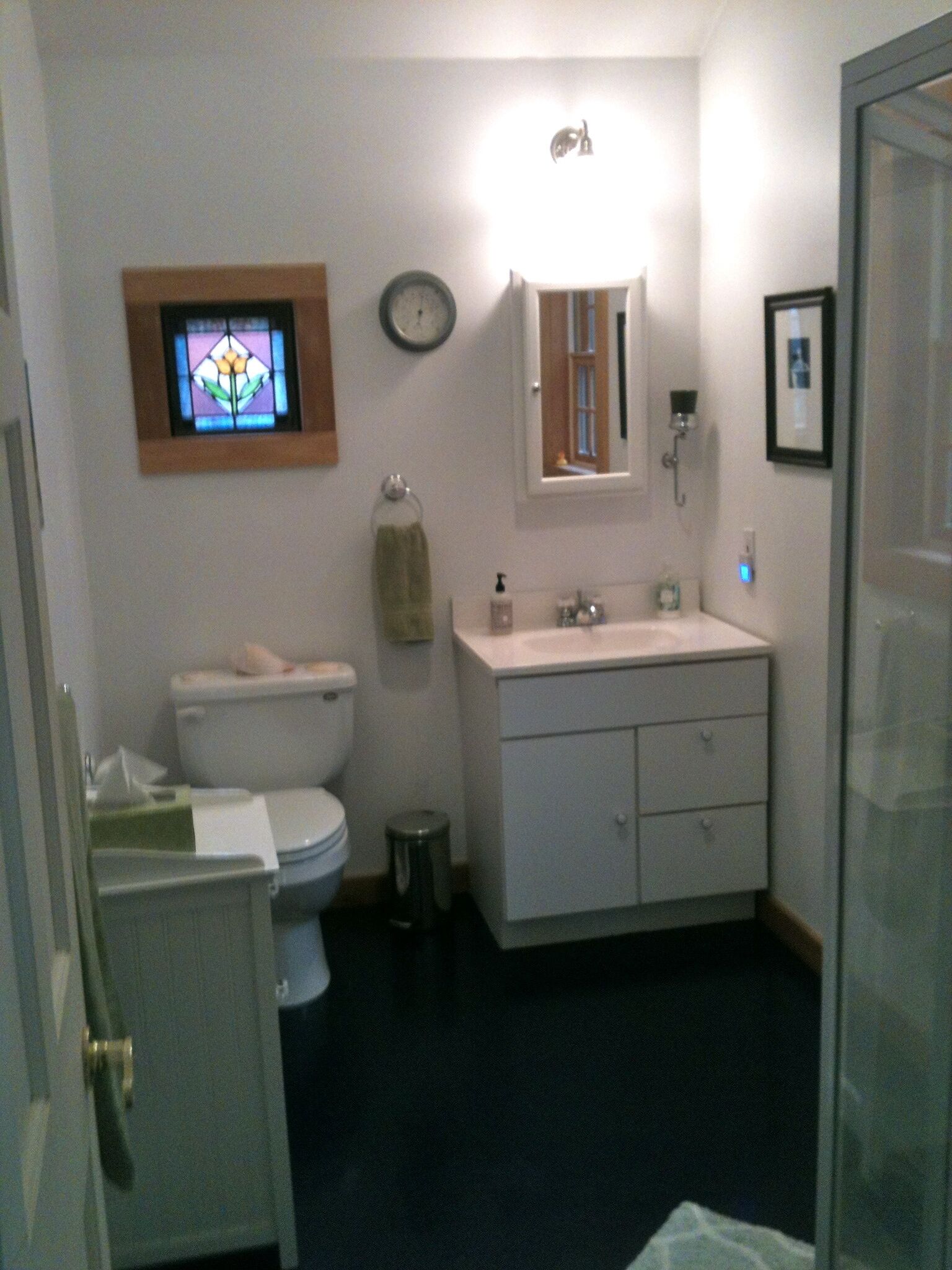 Une salle de bain avec des toilettes, un lavabo et un miroir. Il y a une horloge au mur et un petit tabouret près des toilettes.
