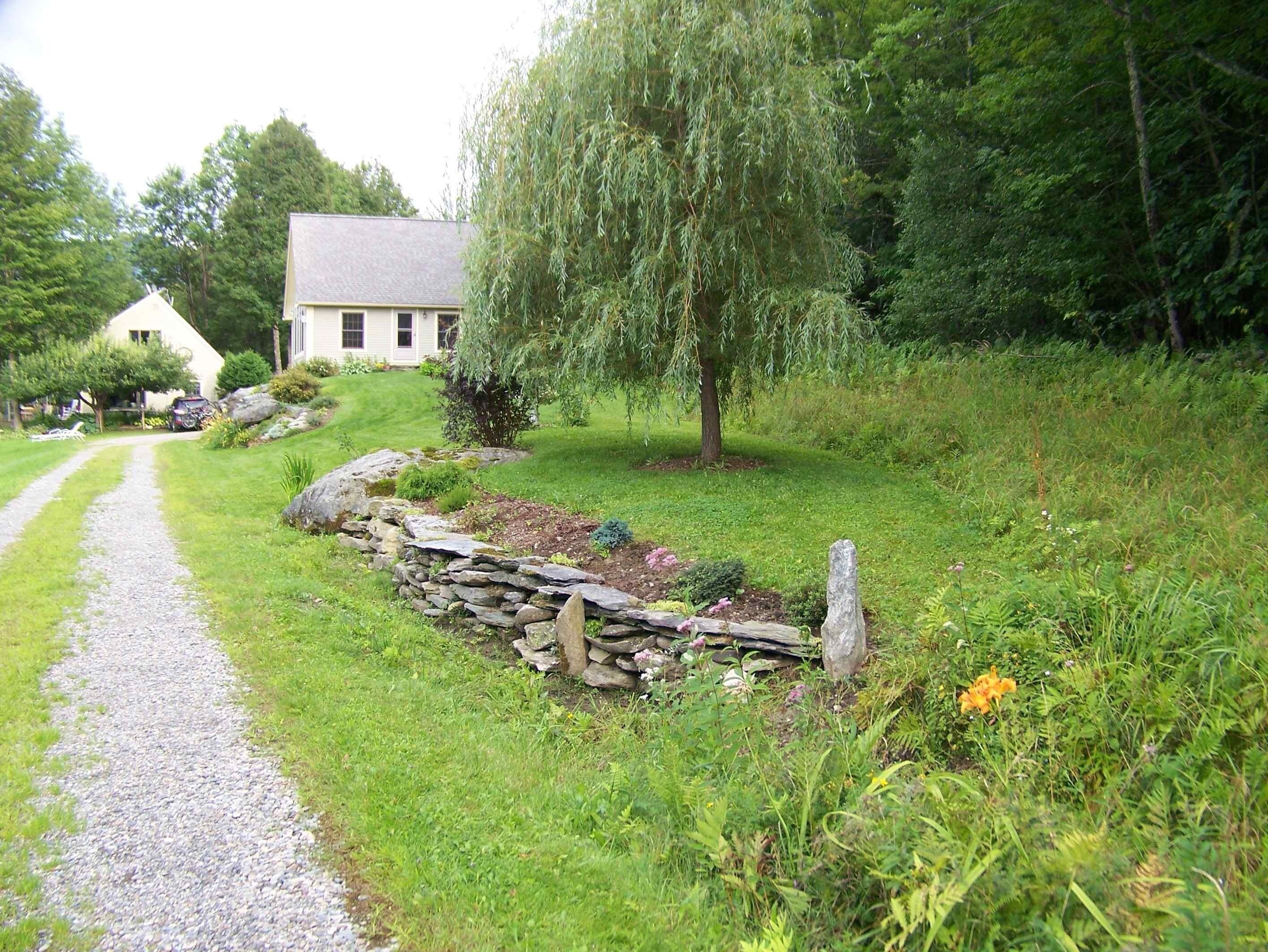 A rural house with a gravel path leading to it, surrounded by lush greenery and a stone wall.