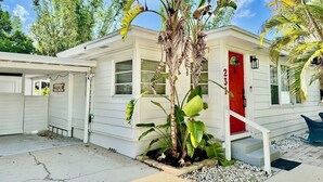 Uma casa térrea com porta vermelha, um pequeno alpendre e uma planta grande no jardim frontal.