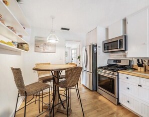 A compact kitchen with white cabinetry, a stainless steel refrigerator, a microwave, and a gas stove. There is a round wooden table with four wicker chairs.