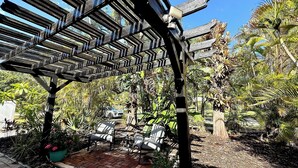 Estrutura de pérgola de madeira com padrão geométrico, rodeada por vegetação exuberante e área pavimentada com assentos externos.