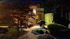 Un jardín bien iluminado con un sendero que conduce a la entrada de un edificio, rodeado de setos cuidados y un árbol con hojas otoñales.