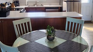 Une cuisine avec des meubles en bois, un réfrigérateur en acier inoxydable, une table à manger avec quatre chaises et une fenêtre avec des stores.