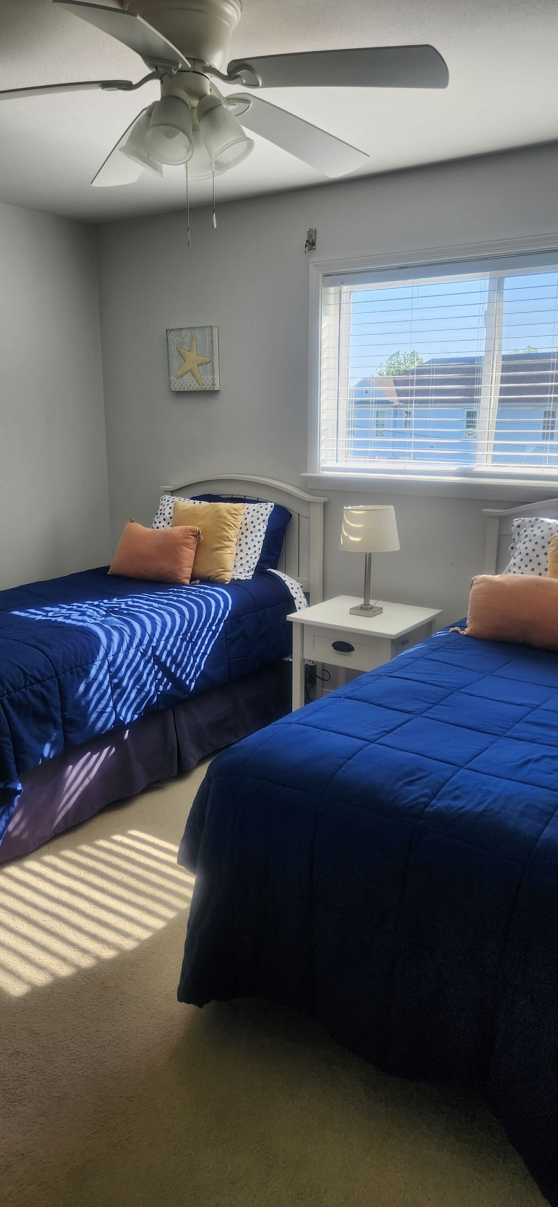 A bedroom with two beds, a ceiling fan, a nightstand, a lamp, and a window with blinds.
