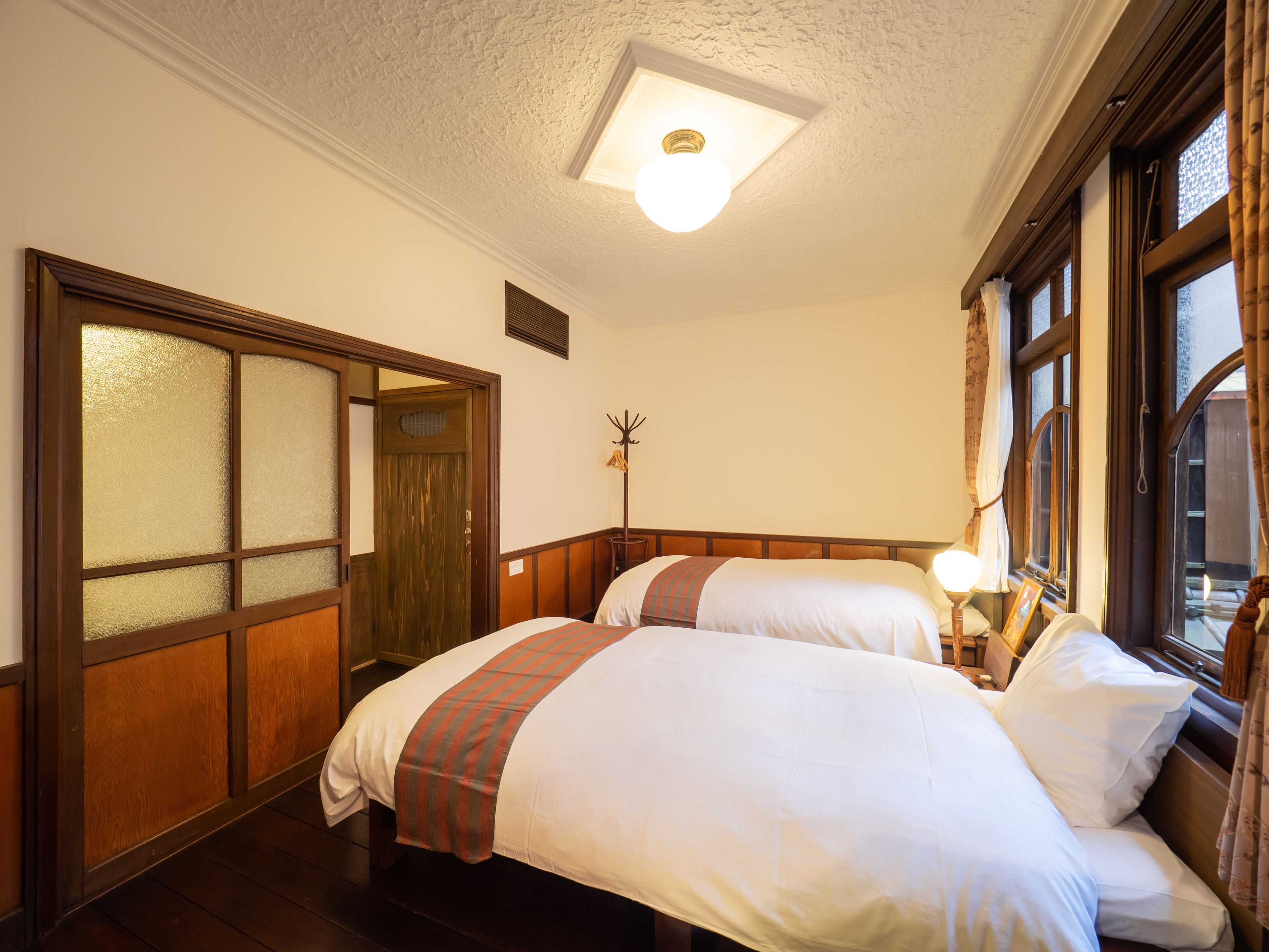 A hotel room with two beds, a wooden door with frosted glass panels, a coat rack, and a window with curtains.