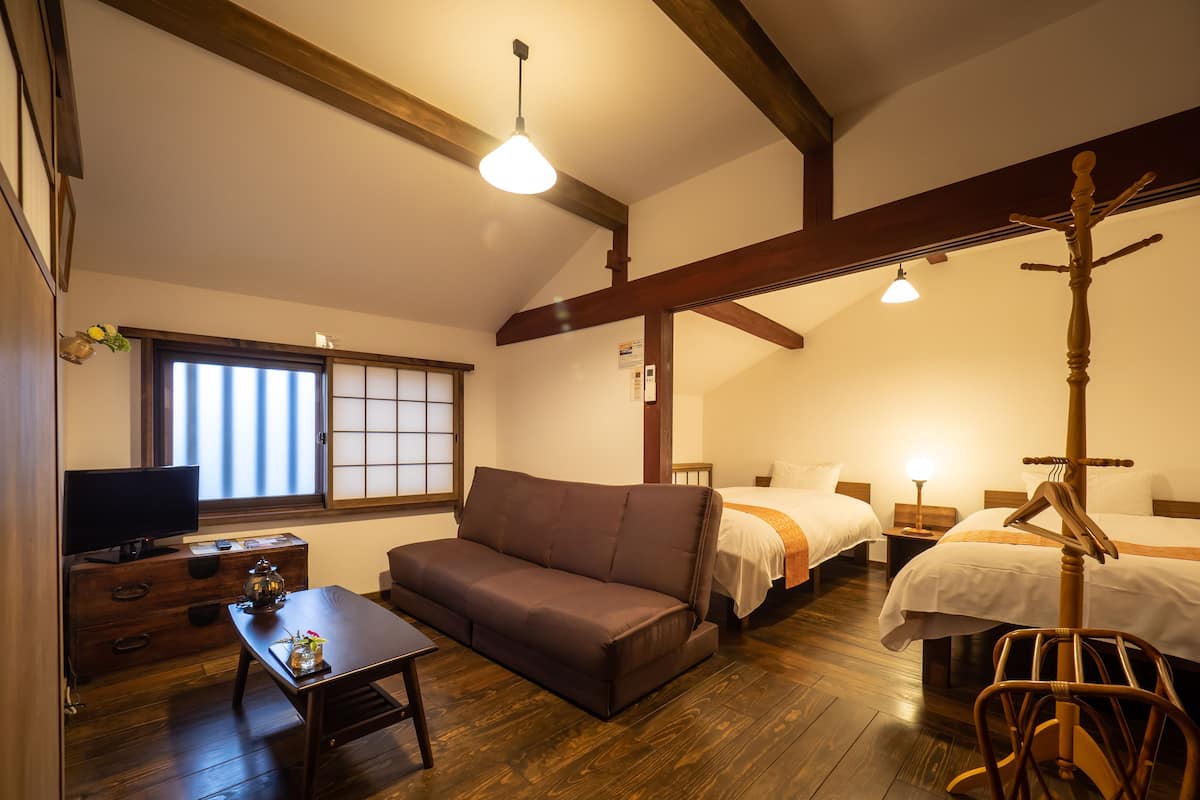 A room with a wooden floor, a sofa, a bed, a nightstand, a television, and a window with blinds.