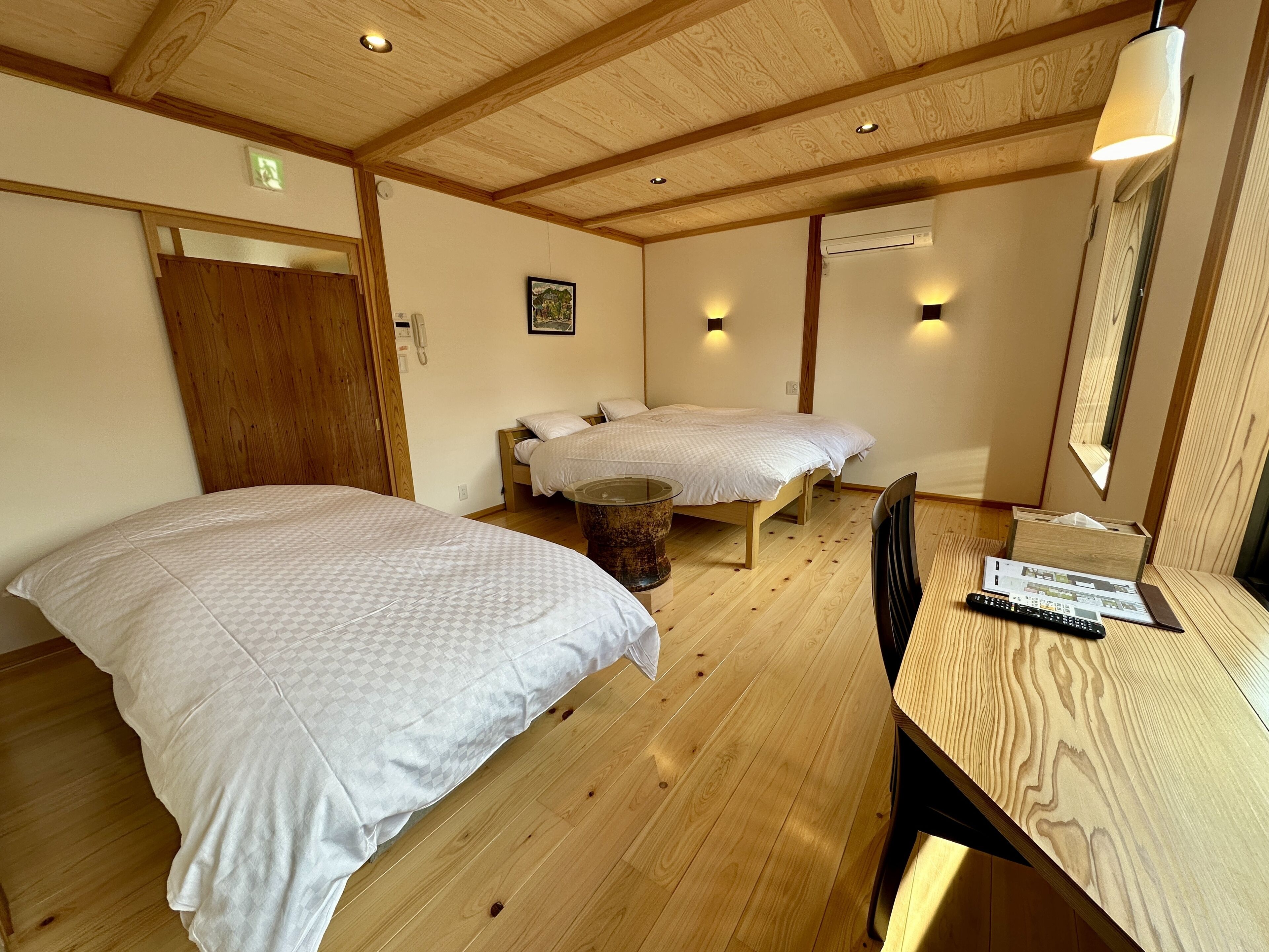 A room with two beds, wooden flooring, a wooden door, a small table with a remote control, and a mirror on the wall.