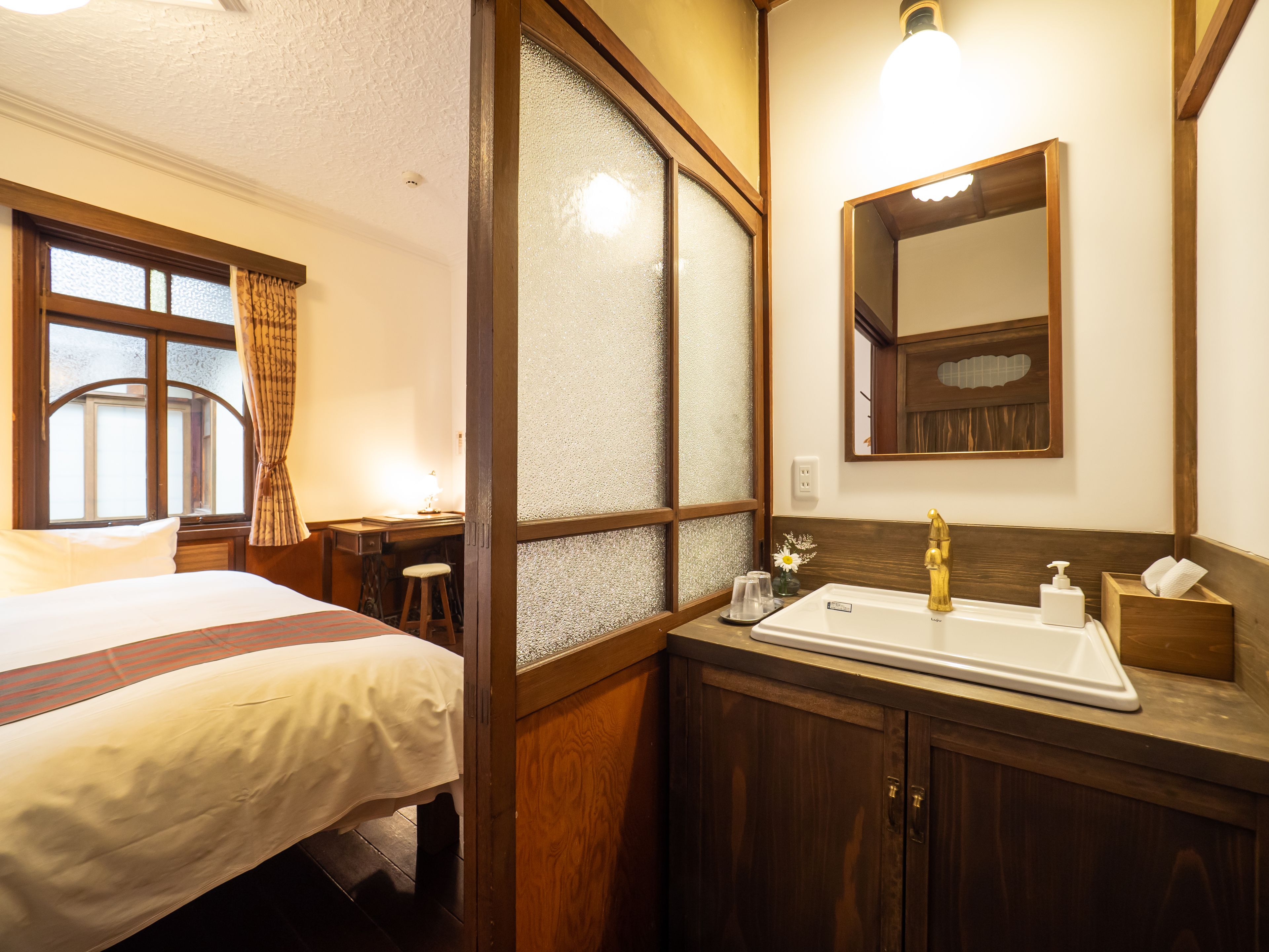 A hotel room with a bed, a desk, a bathroom with a sink and mirror, and frosted glass doors.