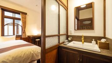 A hotel room with a bed, a desk, a bathroom with a sink and mirror, and frosted glass doors.