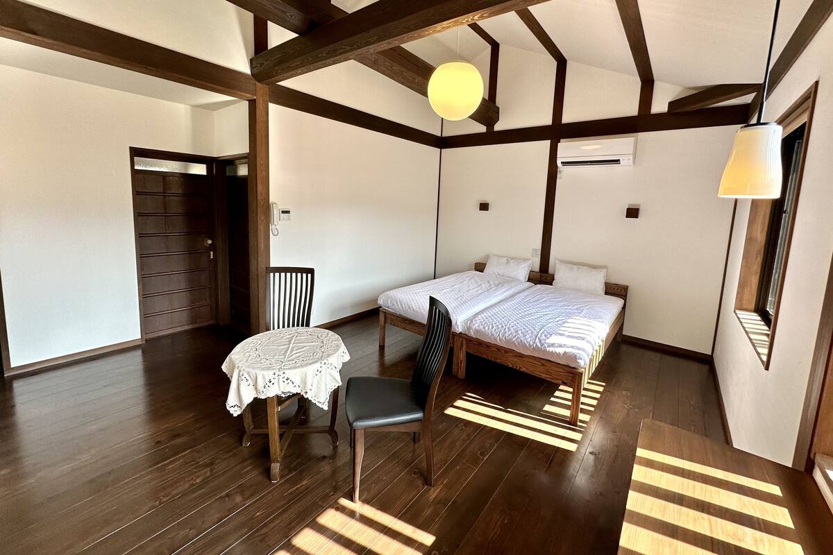 A room with a bed, a table with a lace tablecloth, a chair, and a wooden floor.
