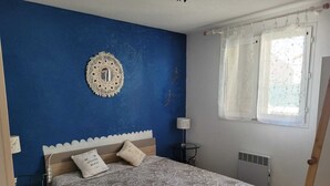 A bedroom with a blue wall, a decorative mirror, a bed with patterned bedding, and a window with curtains. - Marseillan house without opposite in the peace, garden terrace, in a residence (Marseillan)