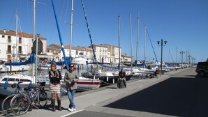 A marina with boats docked, a street with a few pedestrians, and buildings in the background. - Marseillan house without opposite in the peace, garden terrace, in a residence (Marseillan)