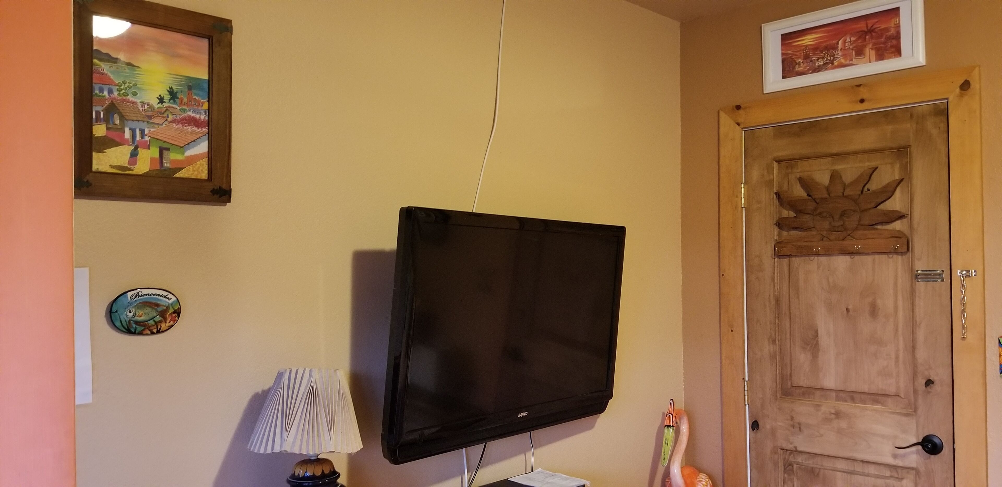 A room with a mounted flat-screen TV, a wooden door with a sun design, and a framed painting on the wall.