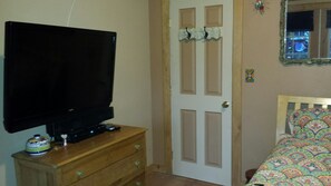 A room with a television on a wooden dresser, a door with a decorative mirror, and a bed with a colorful patterned bedspread.