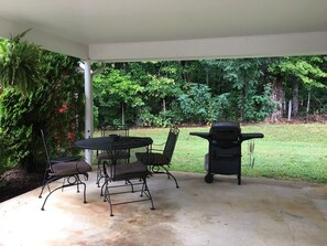 An outdoor patio with a table and chairs, a grill, and a view of a grassy yard with trees.