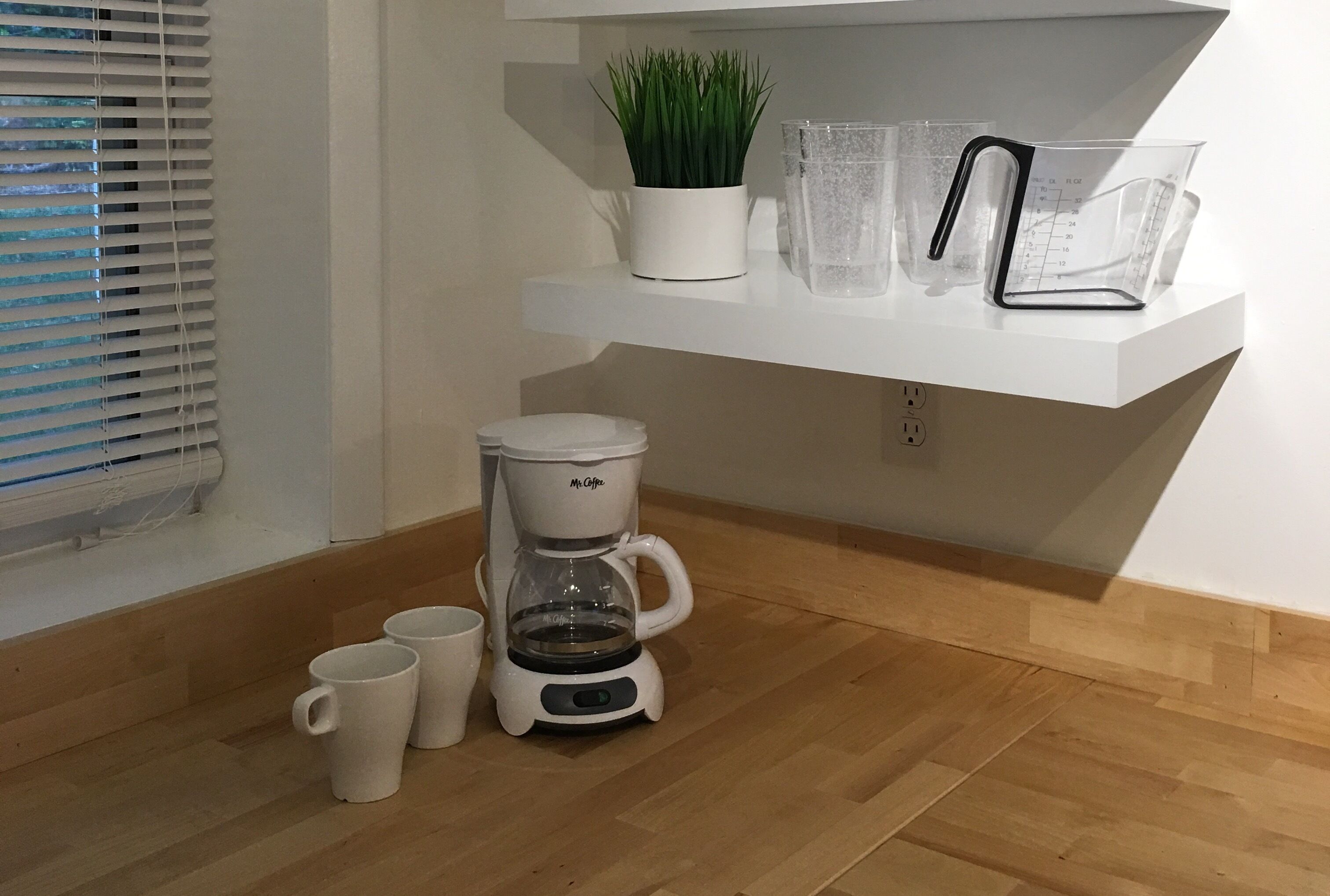 A kitchen area with a coffee maker, two white mugs, a potted plant, and measuring cups on a white shelf.