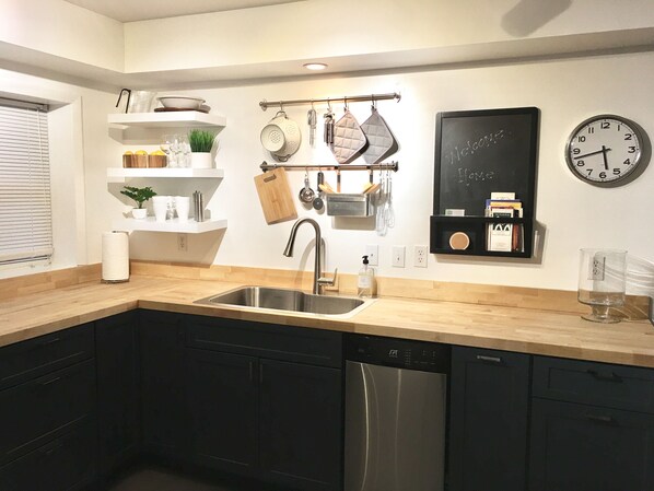 A modern kitchen with a stainless steel sink, black cabinets, and a dishwasher. There are shelves with kitchen items and a chalkboard on the wall.