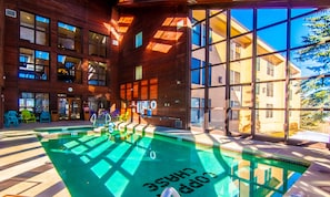 A modern indoor pool with clear glass walls, a wooden exterior, and a view of a neighboring building.