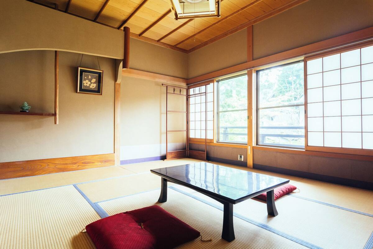 Une pièce avec un sol recouvert de tatamis, une petite table basse, un coussin rouge et un tableau encadré accroché au mur.