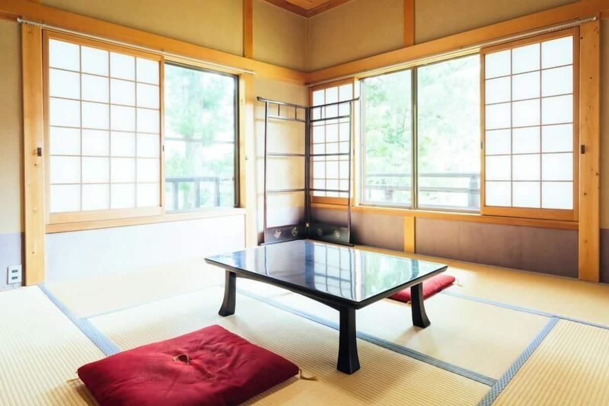 Une pièce avec un sol recouvert de tatamis, une petite table basse, des portes coulissantes et une vue sur la verdure à l’extérieur.
