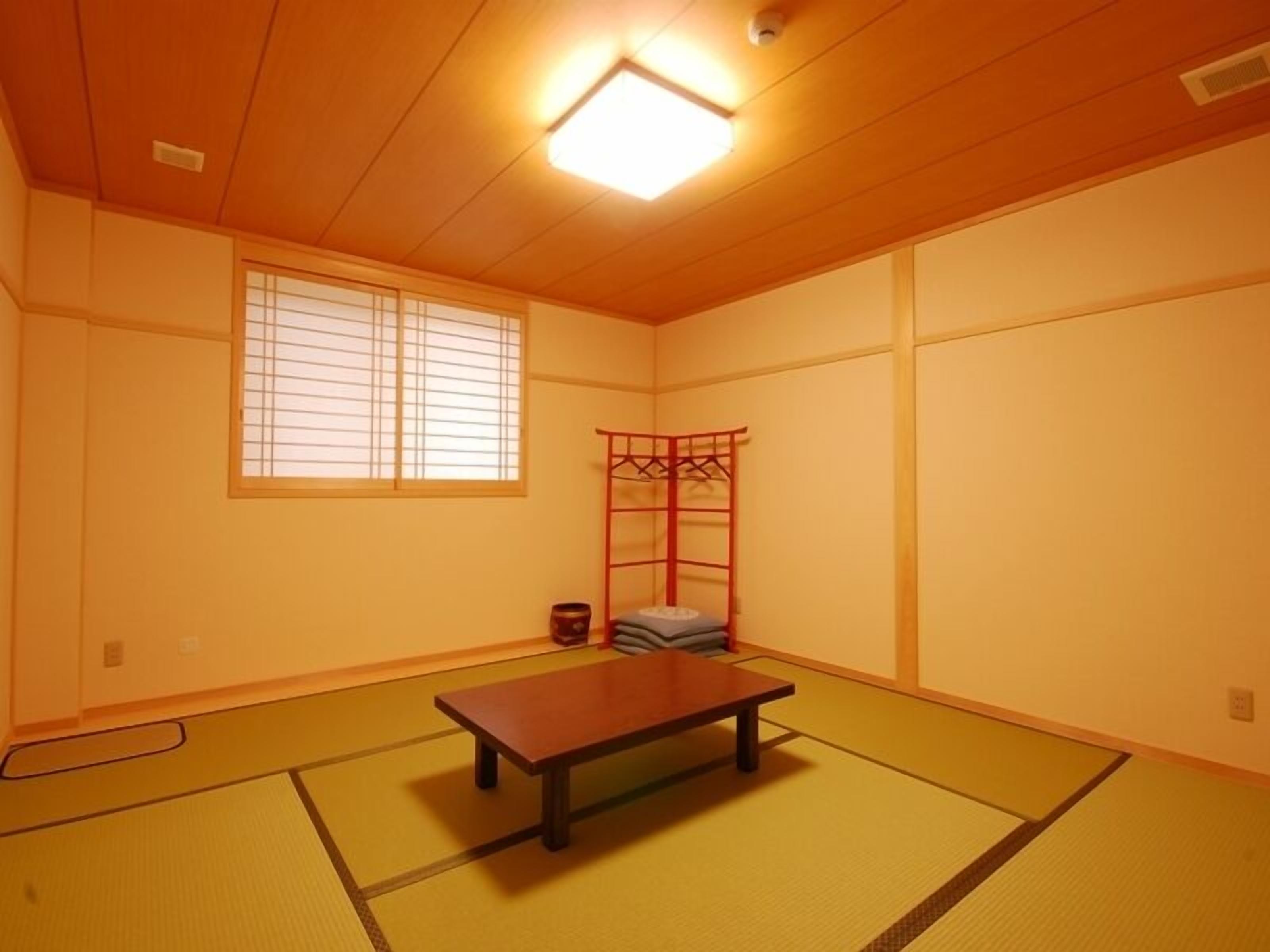 Una habitación con suelo de tatami, una mesa de madera, un mueble de almacenamiento rojo y una ventana con persianas.