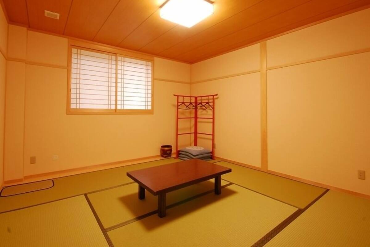Una habitación con suelo de tatami, una mesa de madera, un mueble de almacenamiento rojo y una ventana con persianas.