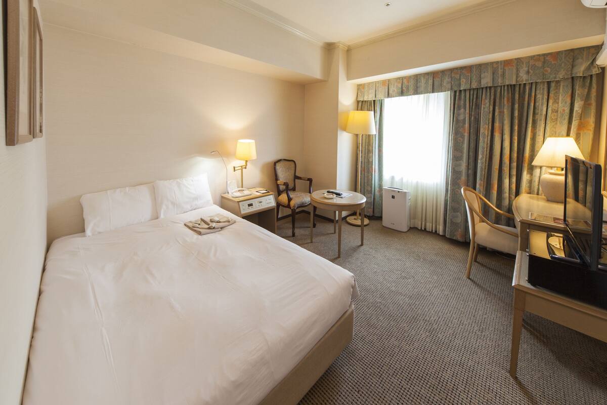 A hotel room with a large bed, a desk with a chair, a small table, and a TV.