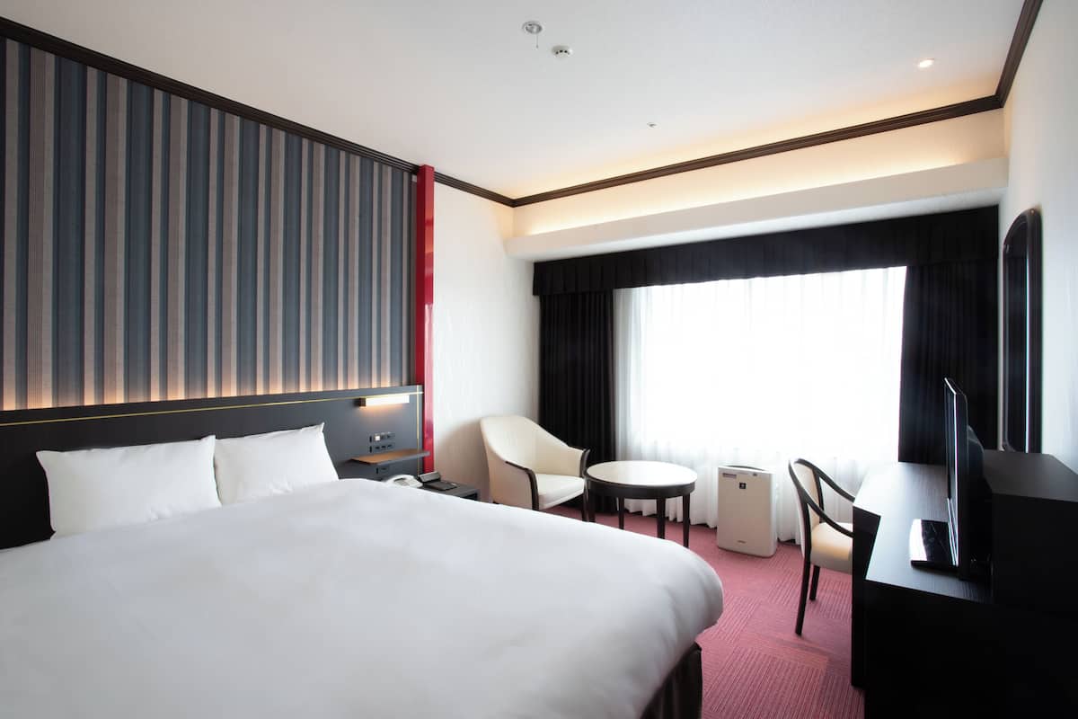 A hotel room with a large bed, a desk with a computer, a chair, a small table, and a window with curtains.