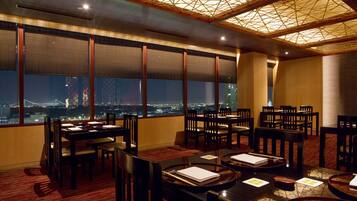 A restaurant with tables set for dining, large windows with a cityscape view, and a ceiling with a geometric pattern.