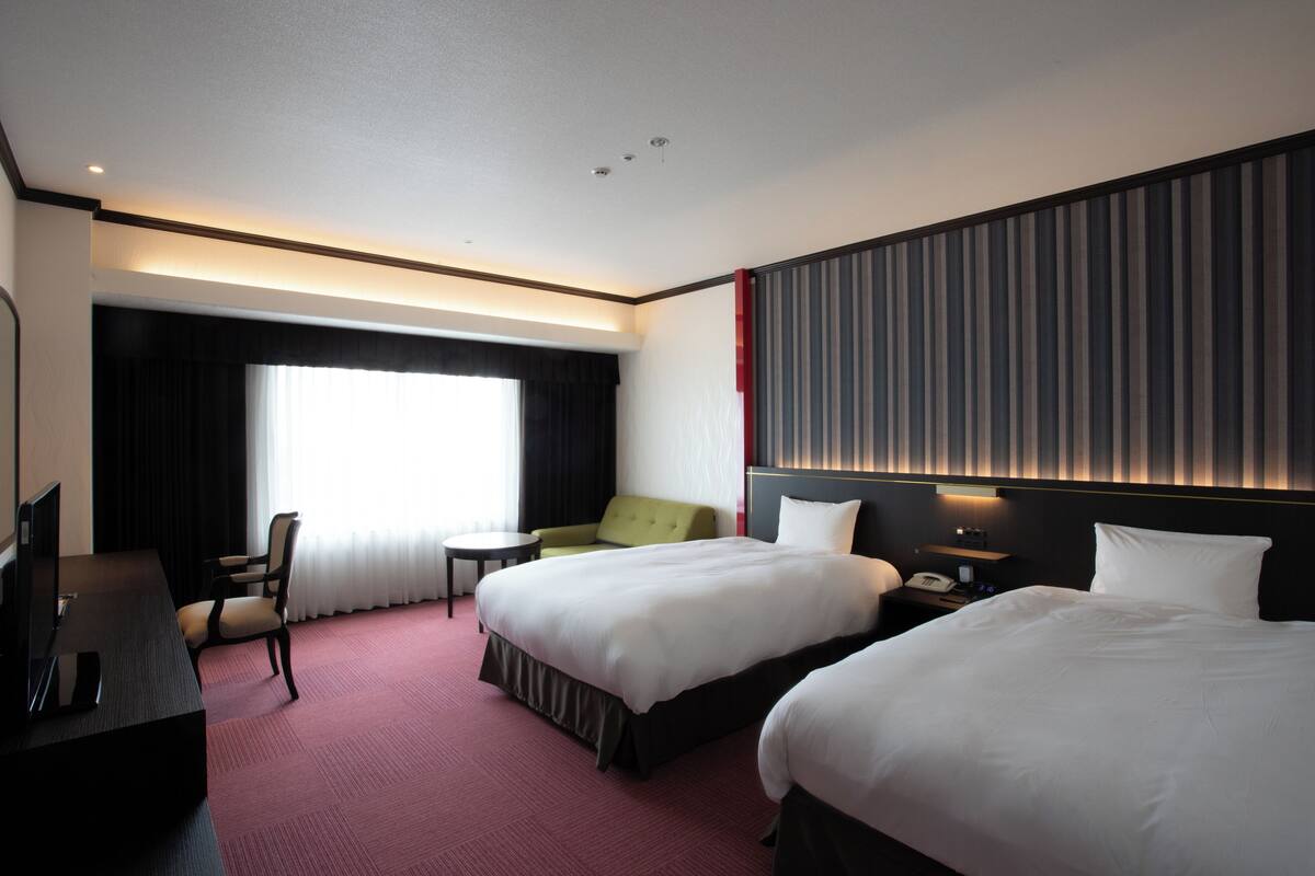 A hotel room with two beds, a desk, a chair, a small table, and a window with curtains.