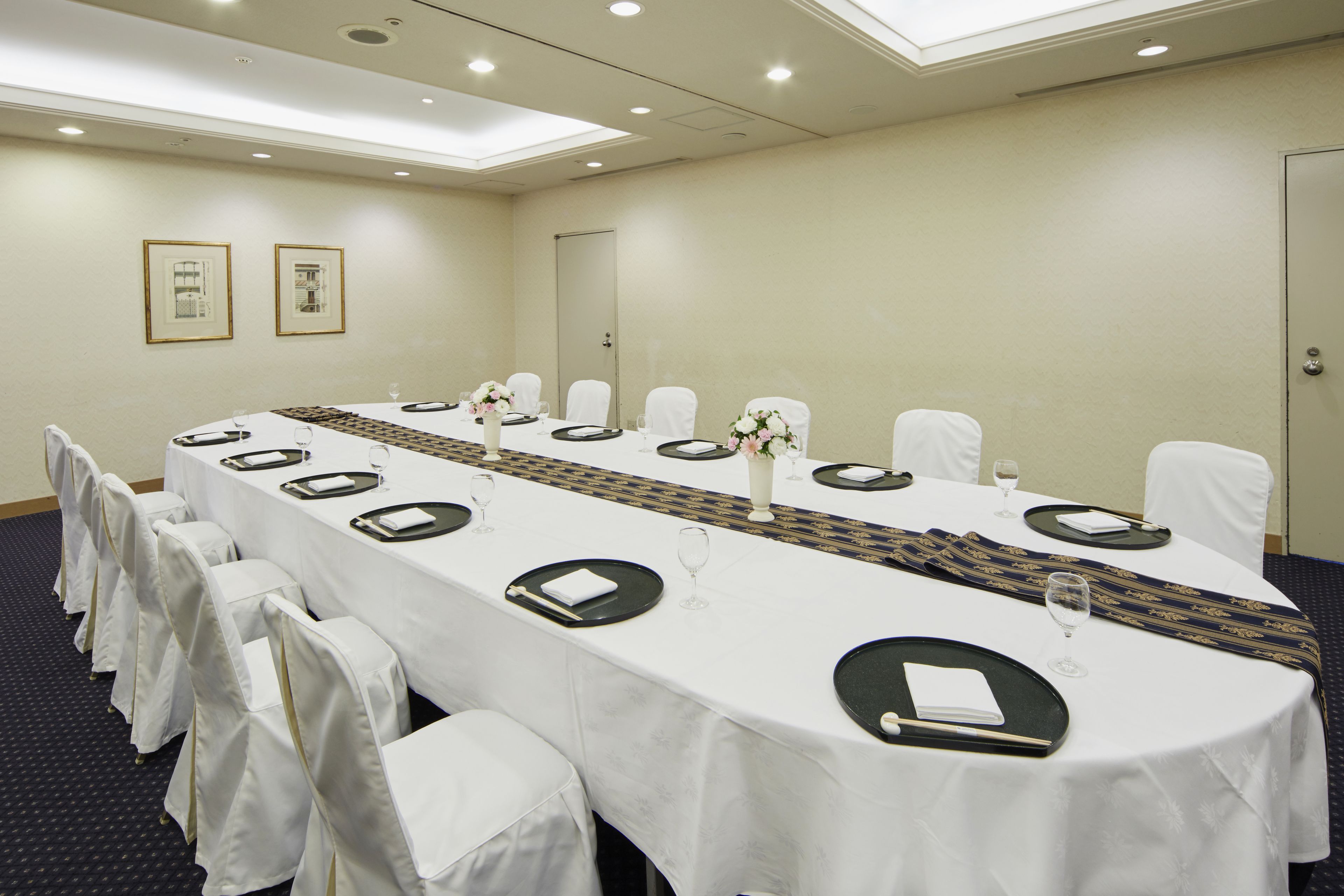 Un salón de conferencias con una larga mesa dispuesta para una reunión, sillas blancas y cuadros en la pared.
