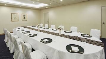 Un salón de conferencias con una larga mesa dispuesta para una reunión, sillas blancas y cuadros en la pared.
