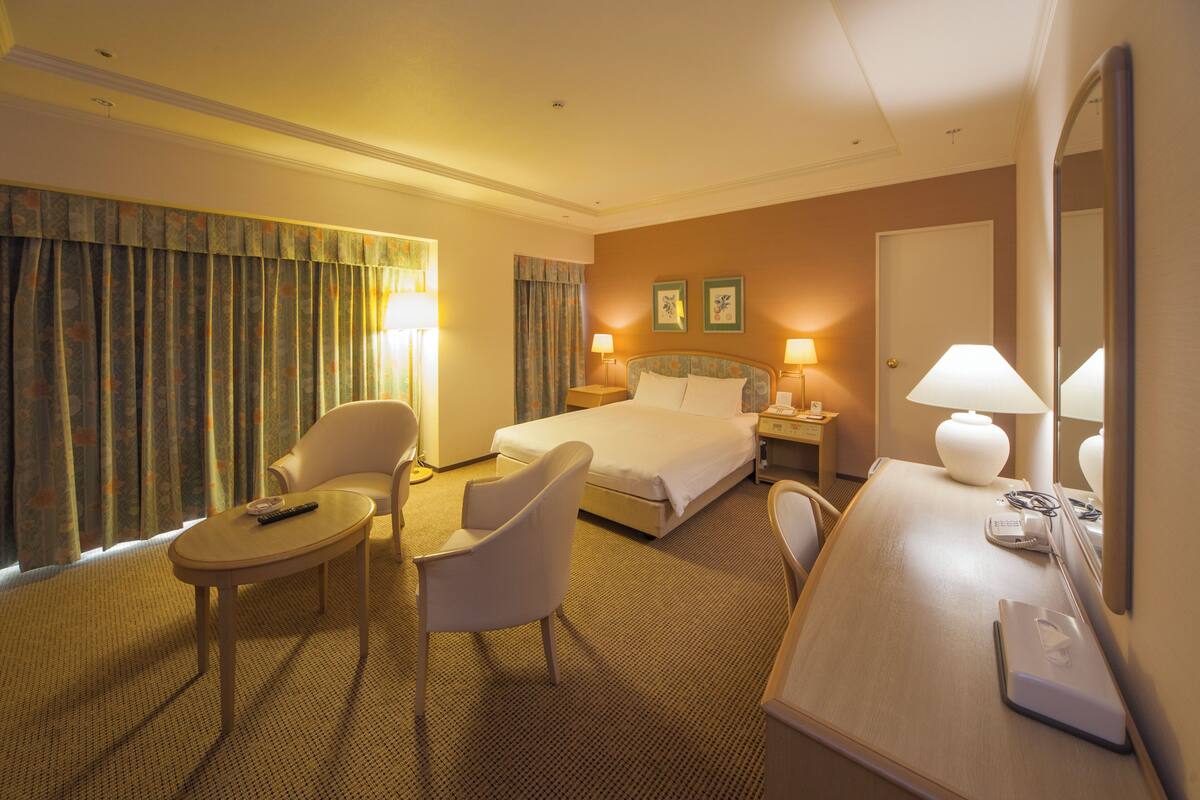 A hotel room with a bed, two chairs, a desk with a lamp, a telephone, and a mirror.