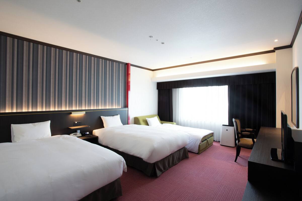 A hotel room with two beds, a desk, a chair, a TV, and a window with curtains.